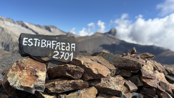 Et toppskilt med teksten ESTIBAFREJA 2701 på en steinvarde, med fjell og blå himmel i bakgrunnen.