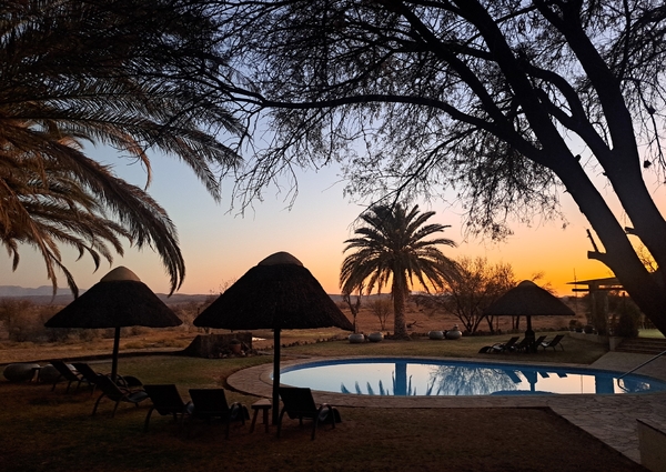 En rolig bassengplass i en afrikansk lodge ved solnedgang, innrammet av silhuetter av trær og stråparasoller mot en fargesprakende himmel.