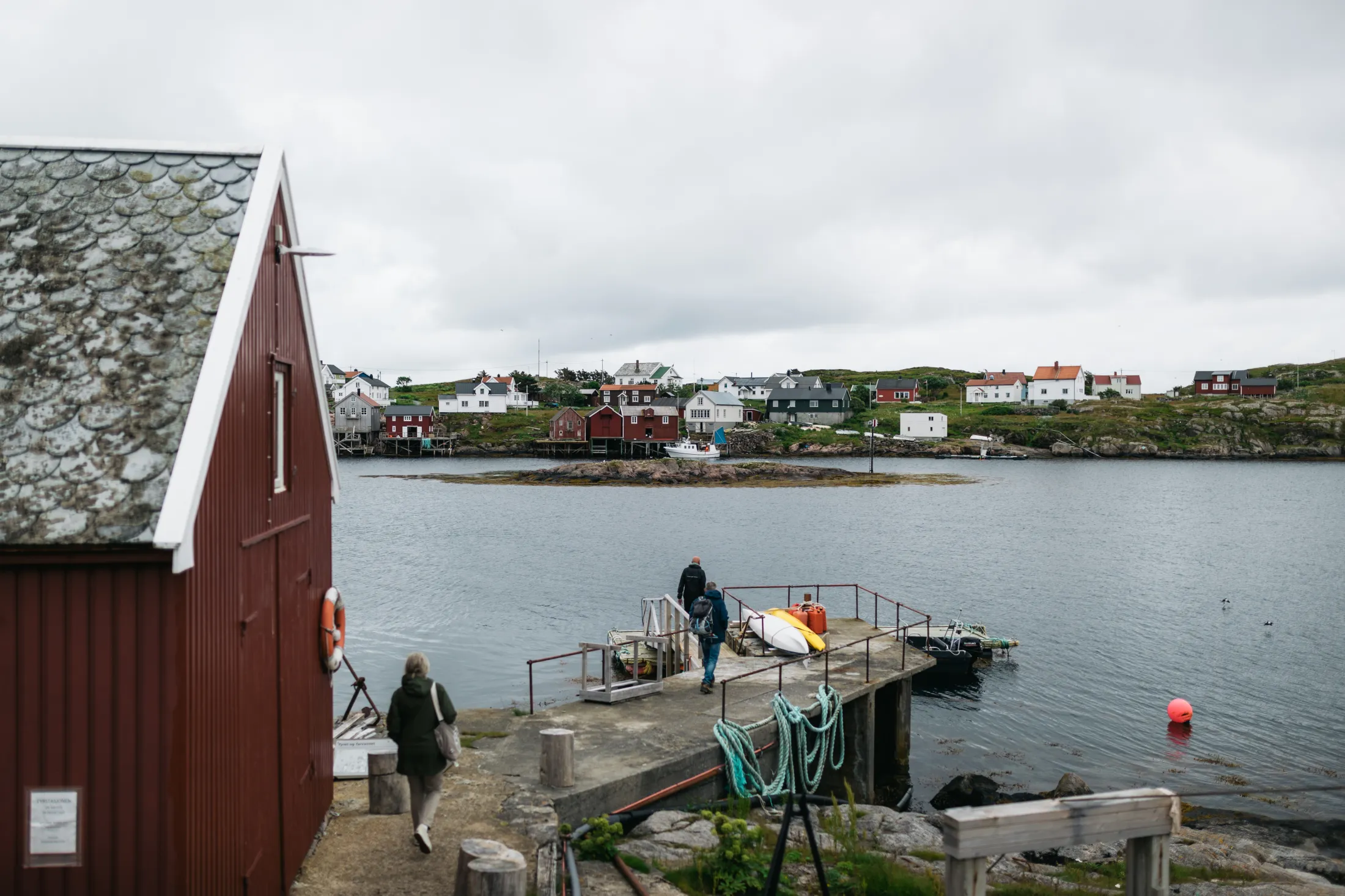Personer som går på en brygge ved et rødt naust, med bebyggelse i bakgrunnen på en overskyet dag.