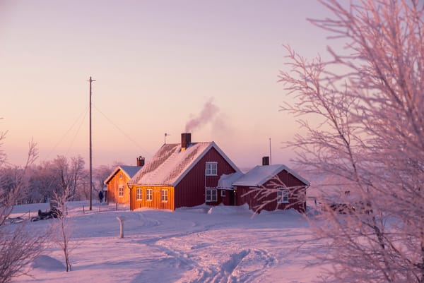 Et koselig rødt hus i et snødekt landskap under en rosa vintersolnedgang, med røyk som stiger opp fra pipa.