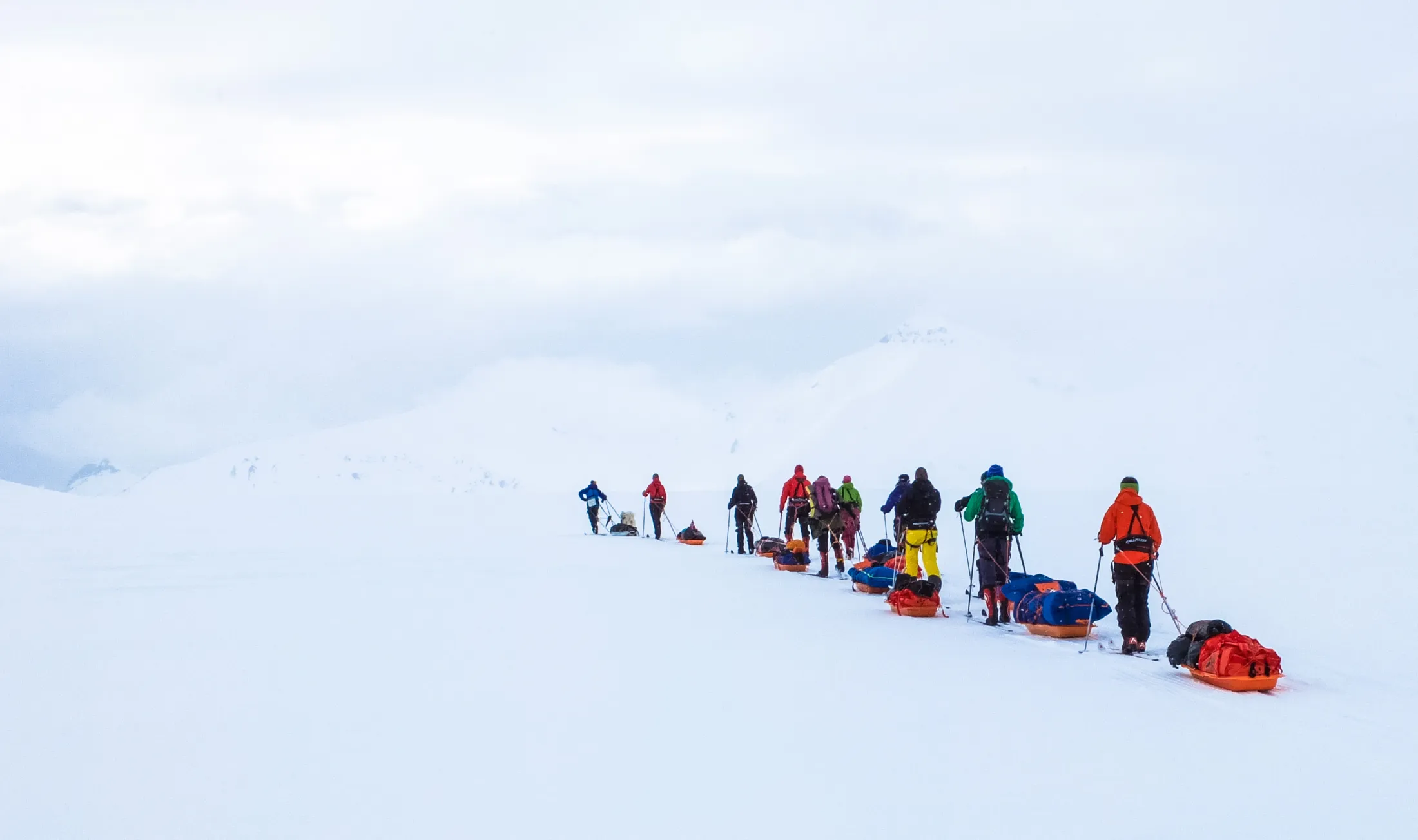 En gruppe skiløpere som trekker pulker beveger seg på rekke over et vidstrakt, snødekt landskap under en overskyet himmel.