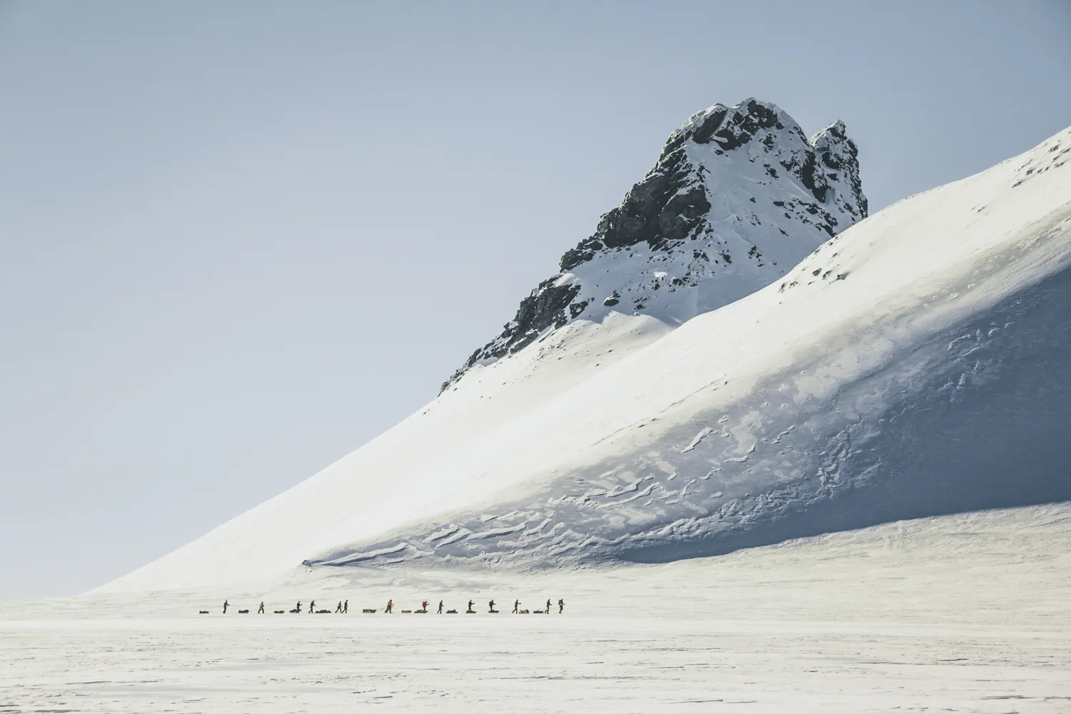 En gruppe mennesker som trekker pulker over et vaidt, snødekt arktisk landskap under en bratt, snødekt fjelltopp.