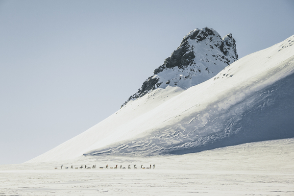 En gruppe mennesker som trekker pulker over et vaidt, snødekt arktisk landskap under en bratt, snødekt fjelltopp.