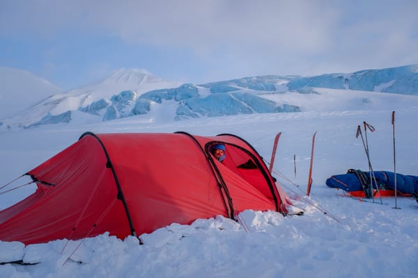 En person titter ut av åpningen på et rødt tunneltelt slått opp i snøen, med skiutstyr stående i nærheten og en enorm, blåtonet isbre og fjell i bakgrunnen under en klar himmel.