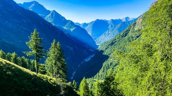 En fantastisk utsikt nedover en dyp fjelldal med en elv som snirkler seg av gårde, rammet inn av bratte, skogkledde grønne skråninger og fjerne blå fjelltopper opplyst av solstråler under en klar himmel.