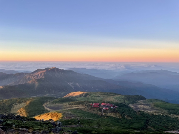 Et fugleperspektiv fanger en klynge med rødtekkede bygninger på et grønt alpint platå under en varm solnedgang som kaster gyldent lys på bølgende fjellrygger under en sart, rosa horisont.