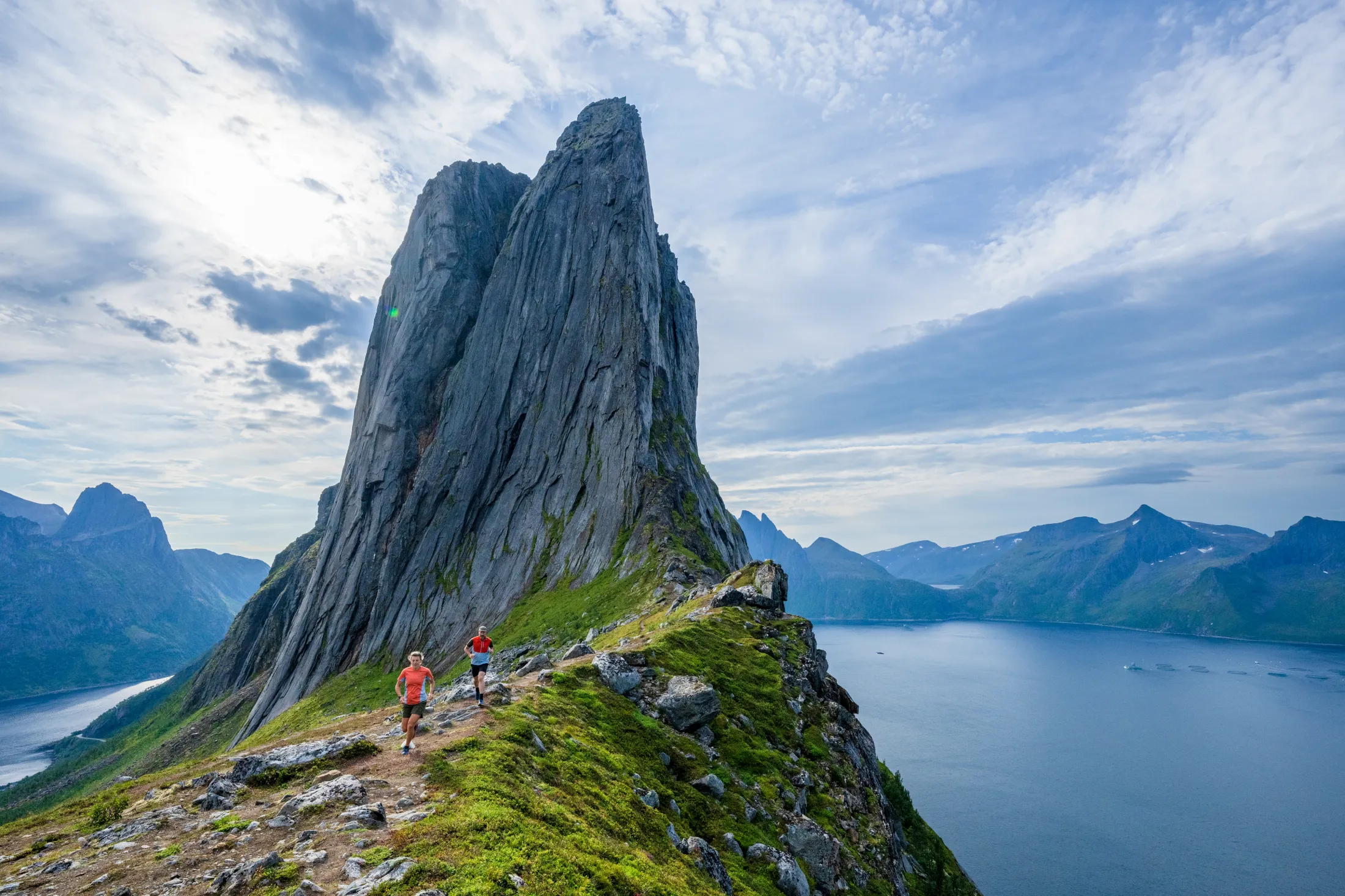 En bratt, dramatisk fjelltopp reiser seg over en fjord med turgåere som går langs en smal rygg i forgrunnen.
