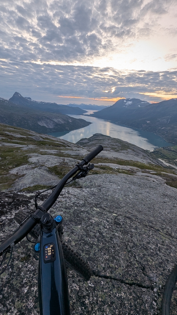 Førstepersonsperspektiv fra en sykkel parkert på en steinete fjellrygg, med utsikt over en bred fjord og fjerne topper under en skyet solnedgangshimmel.