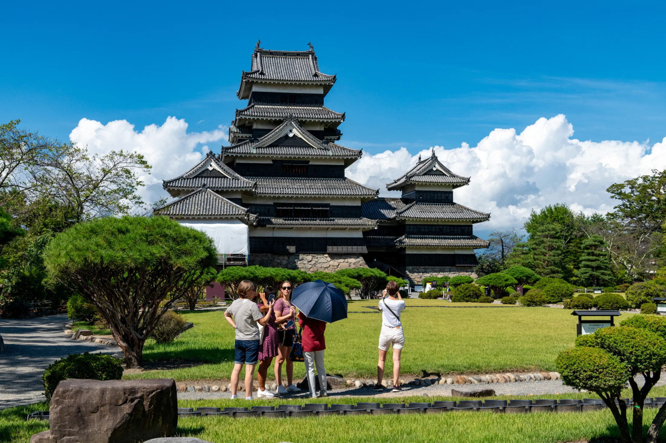 Turister samler seg på en velpleid gressplen for å ta bilder av et storslått, tradisjonelt japansk slott i flere etasjer med mørke trevegger under en klar, blå himmel.