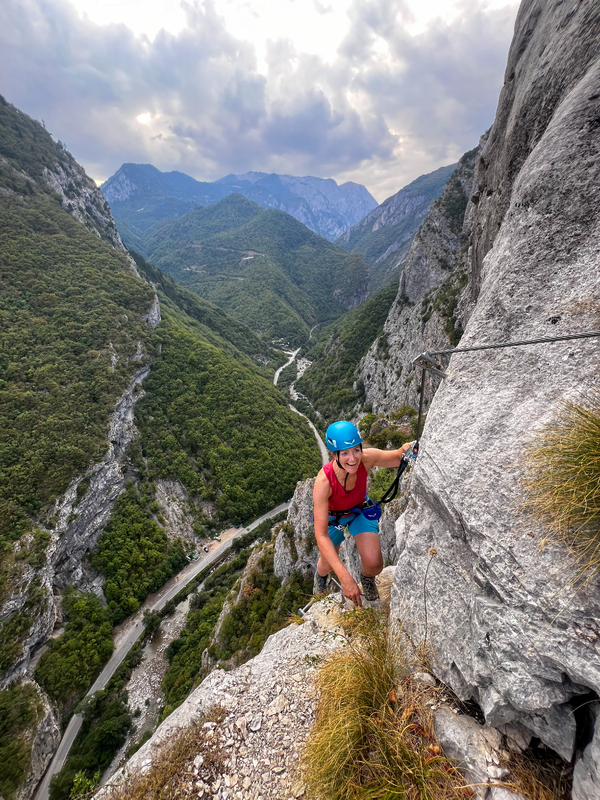 En klatrer på en via ferrata holder i en sikkerhetsvaier med utsikt over en frodig grønn dal med svingete veier og fjerne fjell under en skyet himmel.