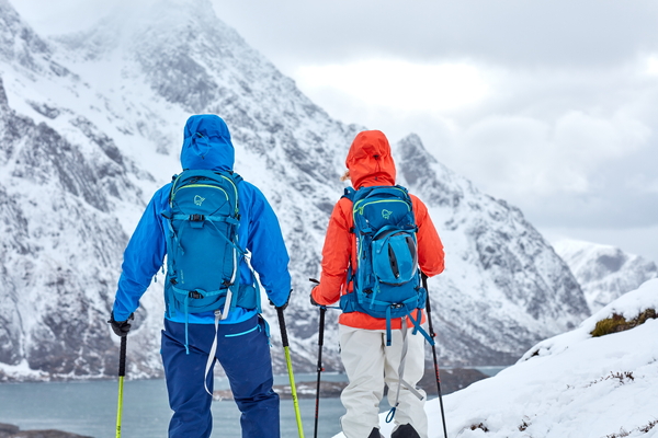 To personer i fargerikt vintertøy og sekker med skistaver, stående i en snødekt fjellside og ser utover et snødekt fjell.