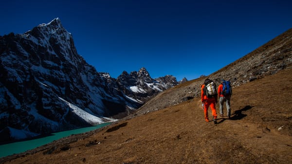 To turgåere med ryggsekker går langs en tørr, gresskledd fjellrygg med utsikt over en turkis bresjø og kneisende, snødryssede fjelltopper under en dyp blå himmel.