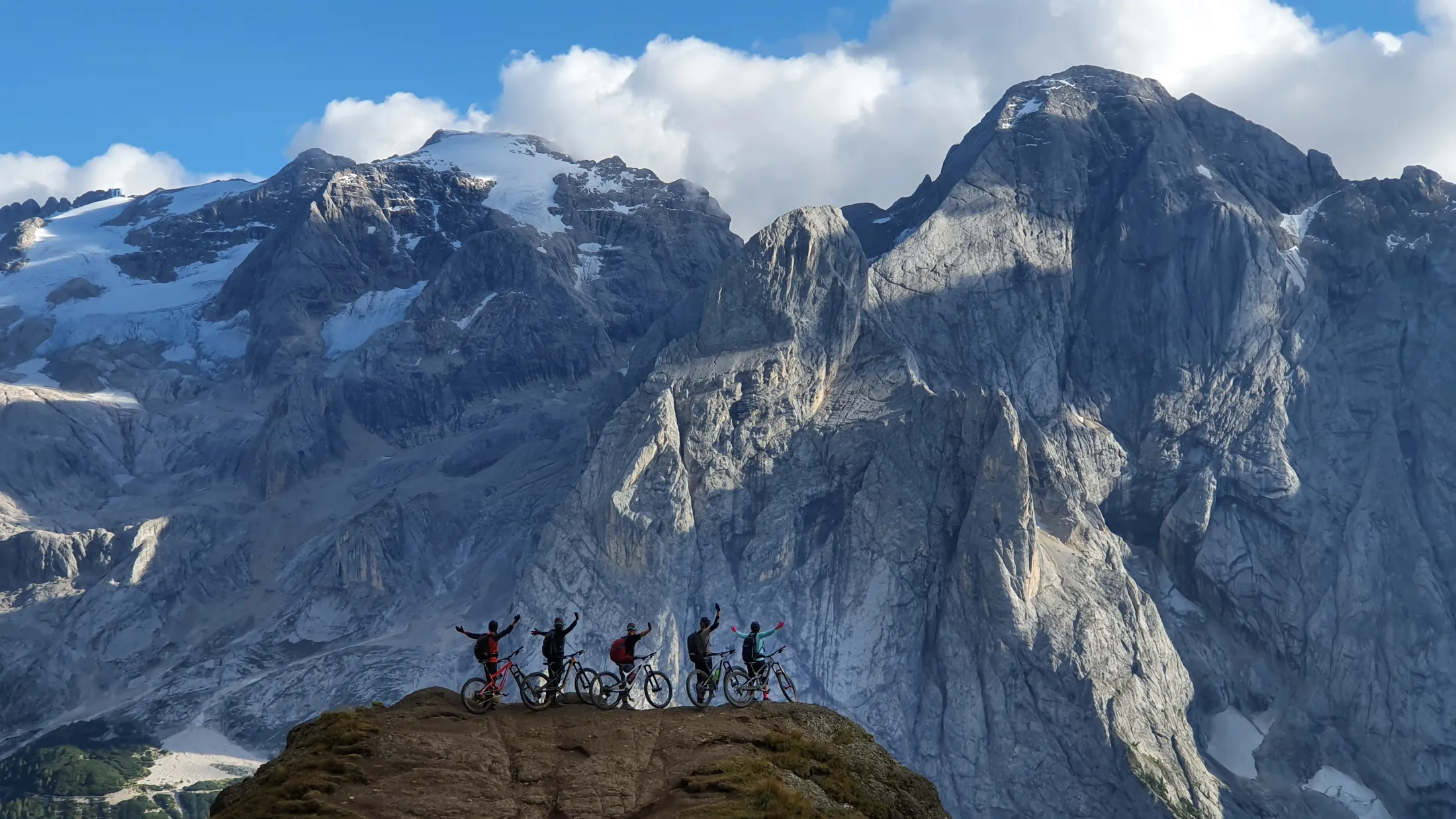 En gruppe turgåere står på en fjelltopp med armene i været, mot et bakteppe av forrevne, snødekte fjell.