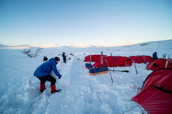 En person som plasserer snøblokker på en levegg ved siden av en rekke med røde ekspedisjonstelt i et snødekt fjellandskap.