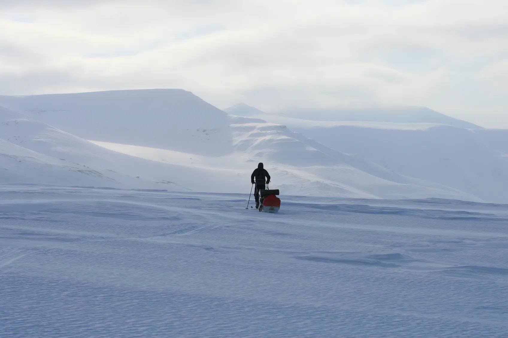 En person som går på ski over et enormt, snødekket arktisk landskap mens de trekker en lastet pulk mot fjerne fjell under en overskyet himmel.
