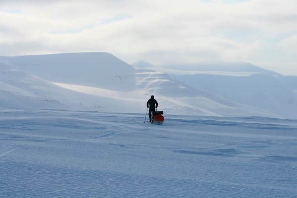 En person som går på ski over et enormt, snødekket arktisk landskap mens de trekker en lastet pulk mot fjerne fjell under en overskyet himmel.