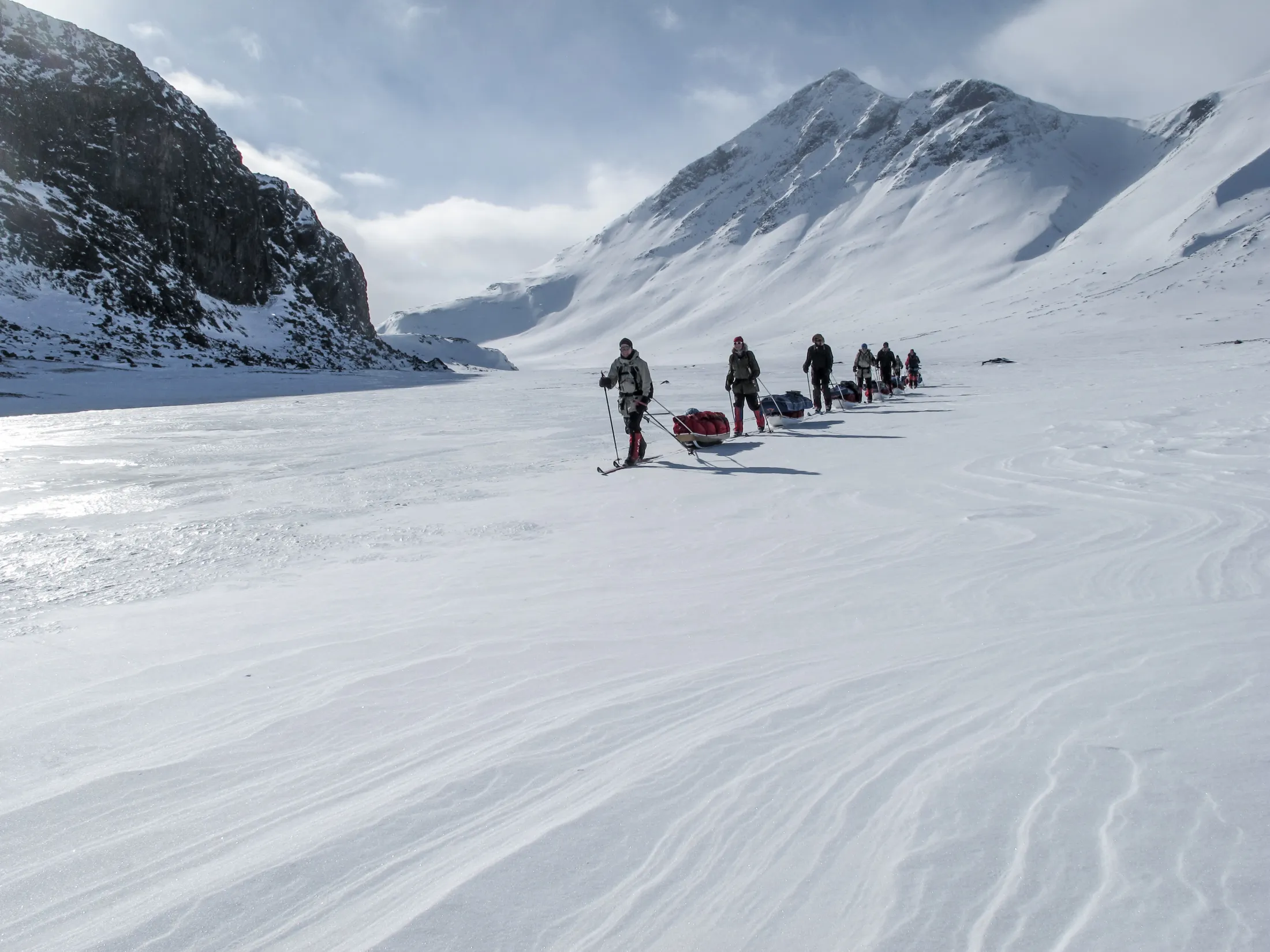 En rekke skiløpere som trekker pulker gjennom en stor, snødekt fjelldal på en solskinnsdag.