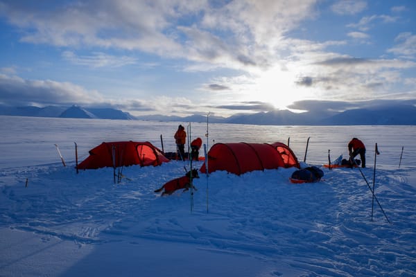 En ekspedisjonsleir med to røde tunneltelt slått opp i snøen, omgitt av mennesker, pulker og en hund i dekken, under en lysende sol som bryter gjennom skyene over en fjern fjellkjede.
