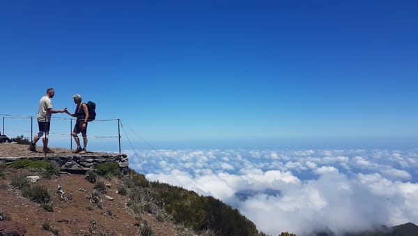 To turgåere som står på et utsiktspunkt i fjellet over et hav av skyer under en blå himmel.