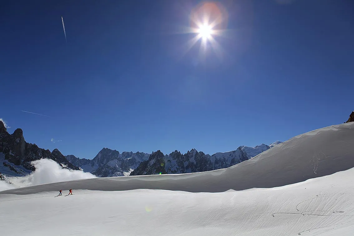 To små skikkelser går på ski over en enorm, snødekt bre under en intens sol og en strålende blå himmel rammet inn av spisse alpine tinder.
