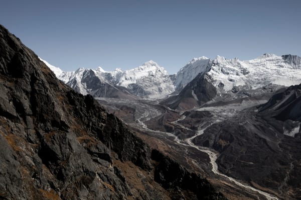 Sett forbi en bratt, mørk og steinete fjellside i forgrunnen, strekker en bred bredal seg ut nedenfor og leder mot en dramatisk bakgrunn av massive, snødekte himalayatopper under en lyseblå himmel.