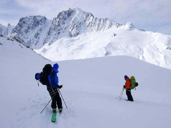 To toppturvandrere med store ryggsekker og skistaver krysser et snødekt fjellandskap under en lys himmel med dramatiske tinder i bakgrunnen.