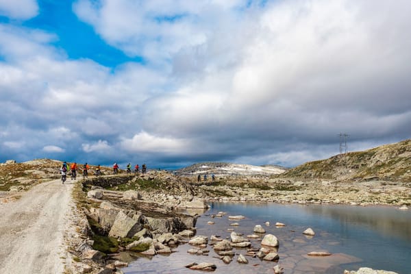 En gruppe syklister sykler langs en steinete fjellsti ved siden av et klart, steinfylt fjellvann under en dramatisk, skyet himmel.