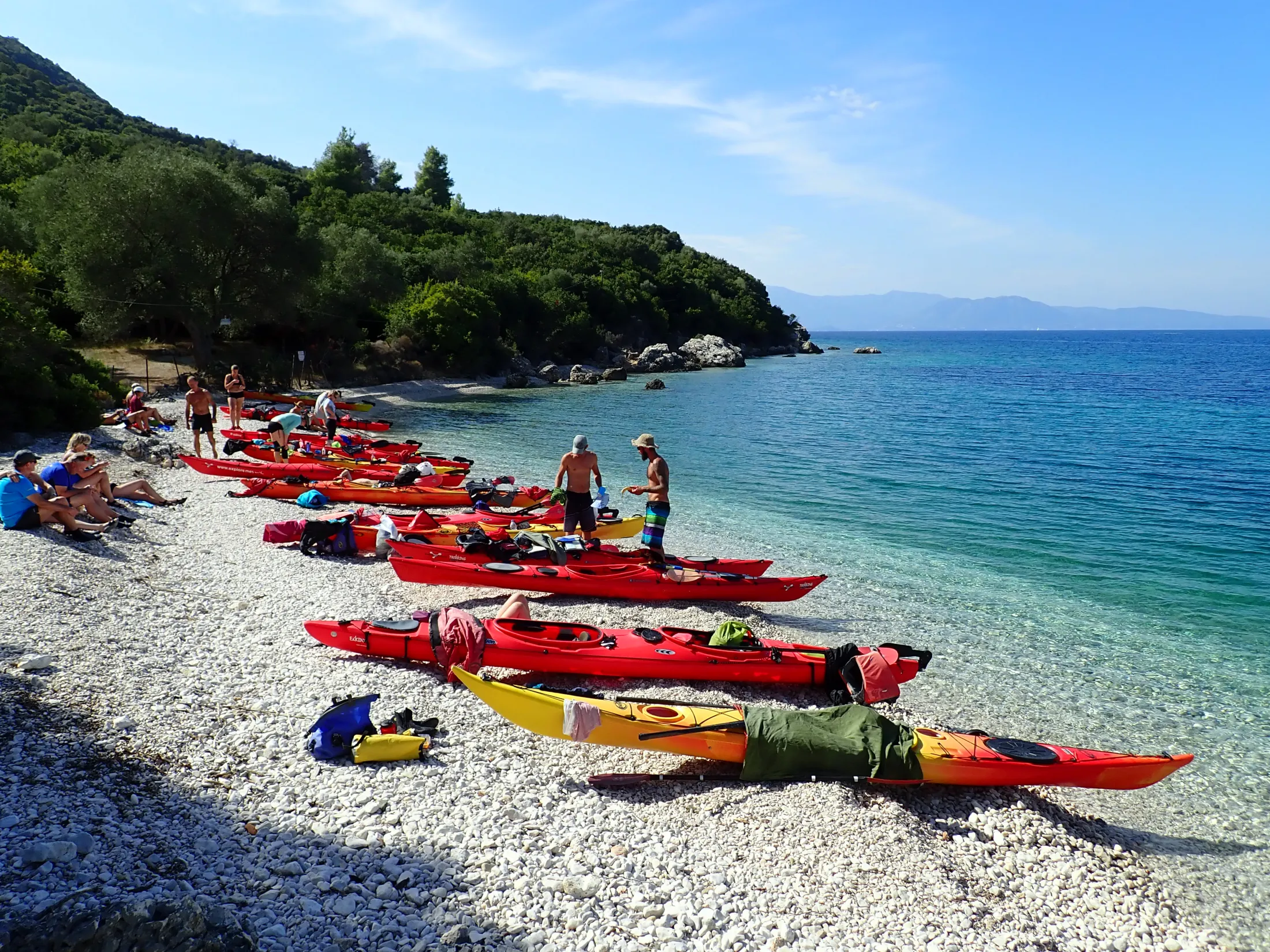 En gruppe mennesker gjør klar sine røde og gule havkayakker langs en hvit rullesteinstrand omkranset av klart turkisblått vann og frodige, grønne åssider.