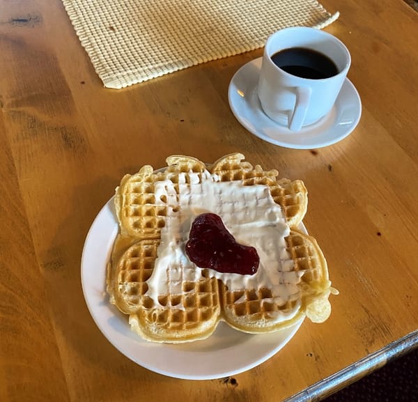 En norsk hjertevaffel med rømme og syltetøy, og en kopp svart kaffe på et trebord.