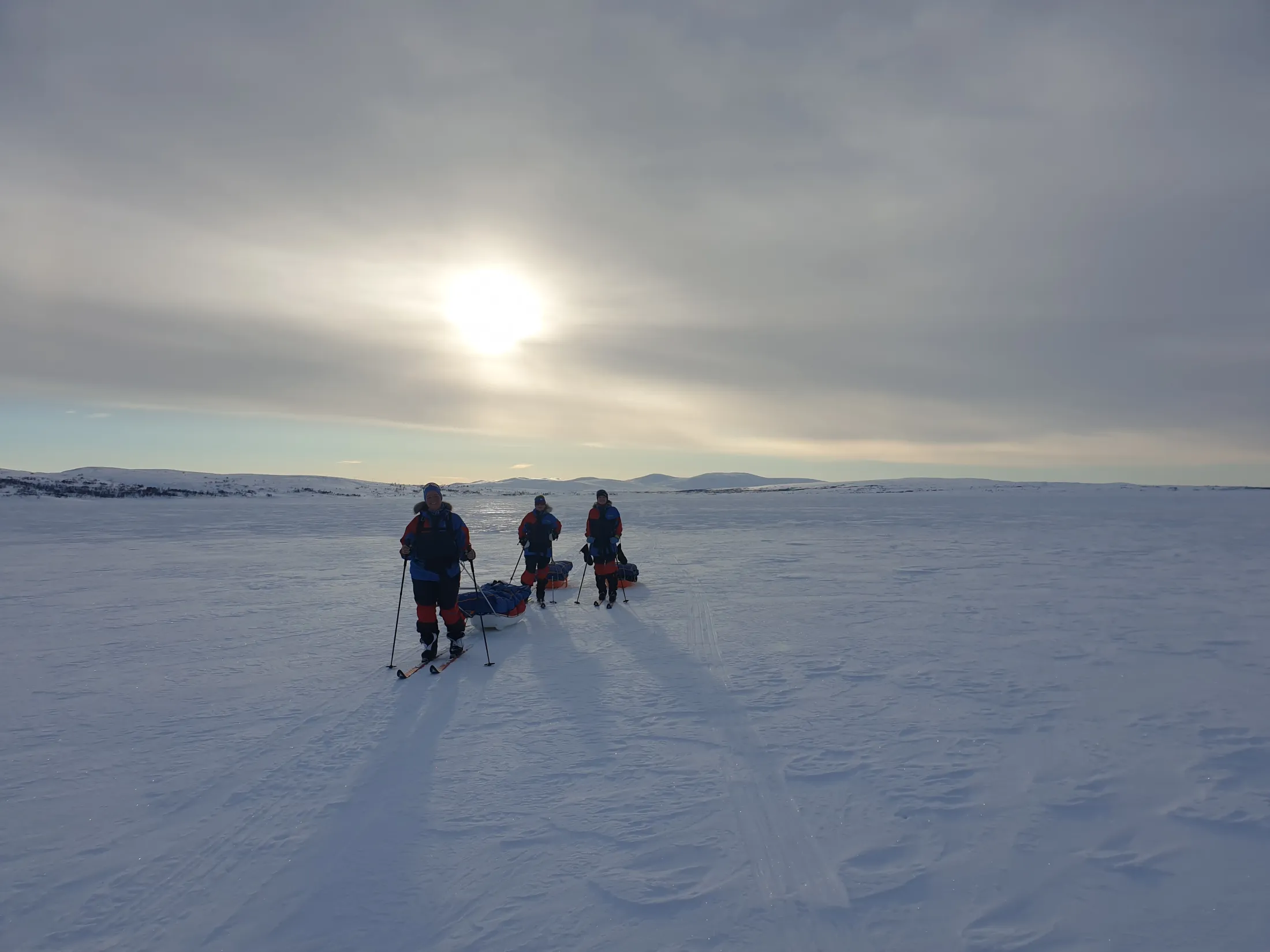 Tre personer krysser et stort snølandskap på ski, og trekker pulker under en skyet himmel der solen skinner igjennom.