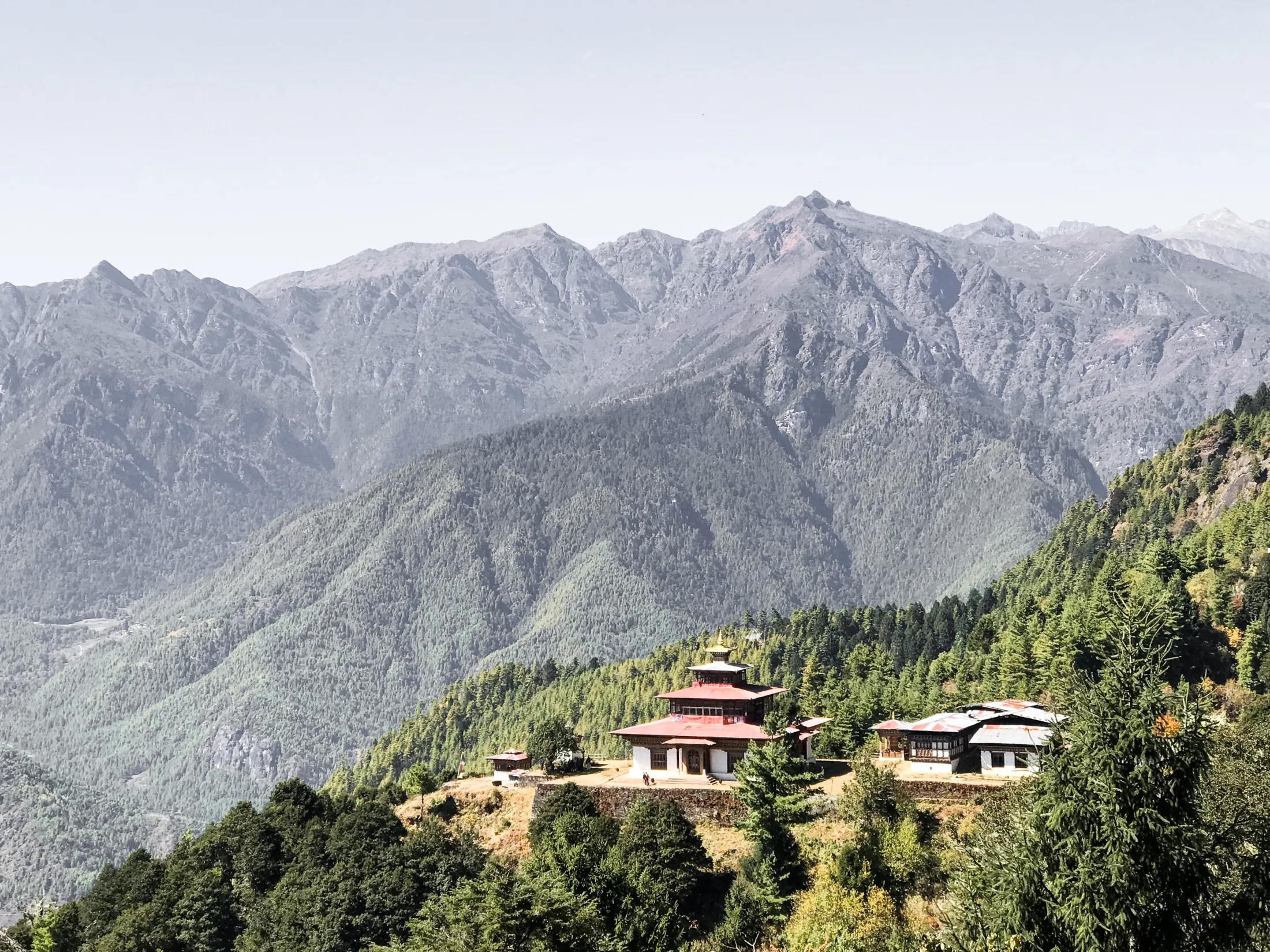 Tradisjonelt bhutansk tempel beliggende på en furukledd åsrygg med mektige fjellkjeder i bakgrunnen.