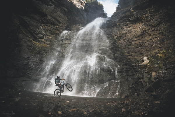 En terrengsyklist som utfører et stunt foran en majestetisk foss som renner nedover en steinete fjellvegg.