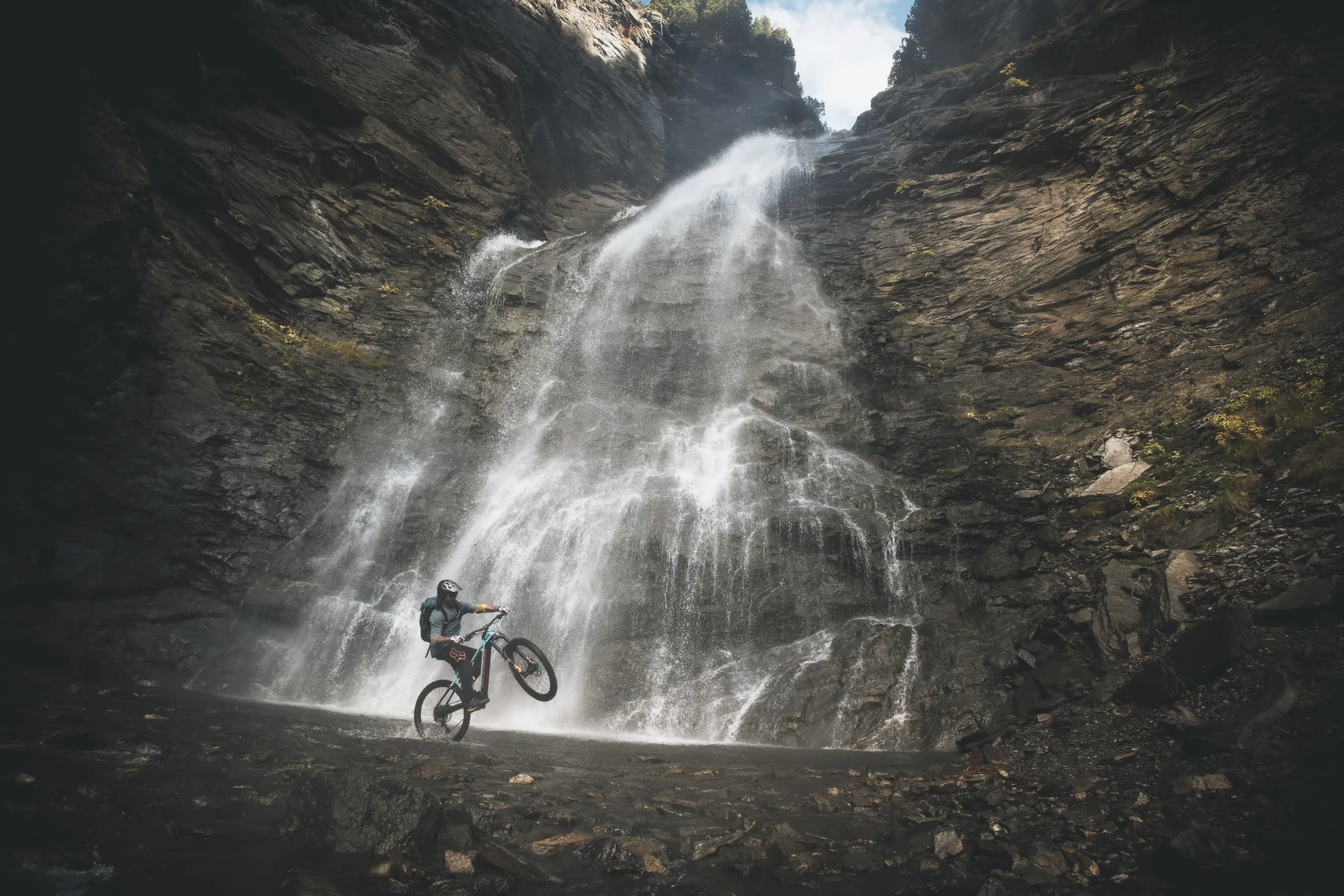En terrengsyklist som utfører et stunt foran en majestetisk foss som renner nedover en steinete fjellvegg.