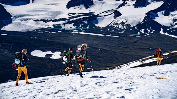 Fjellvandrere går opp en snødekt fjellskråning med et stort brelandskap i bakgrunnen.