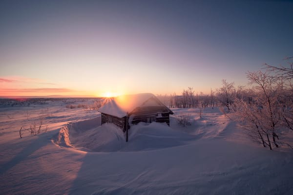 En tømmerhytte omgitt av dyp snø under en fargerik soloppgang.
