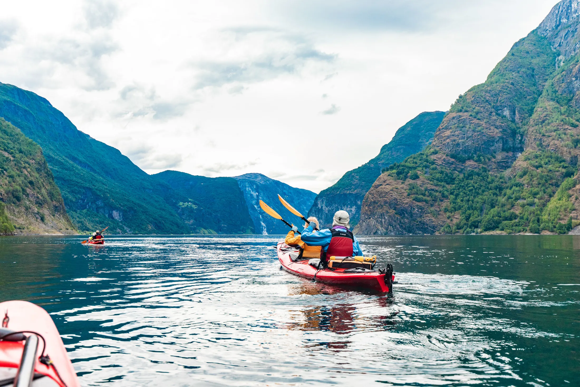 To personer padler i en rød tandemkajakk på en rolig fjord omgitt av høye, frodige, grønne fjell under en overskyet himmel.