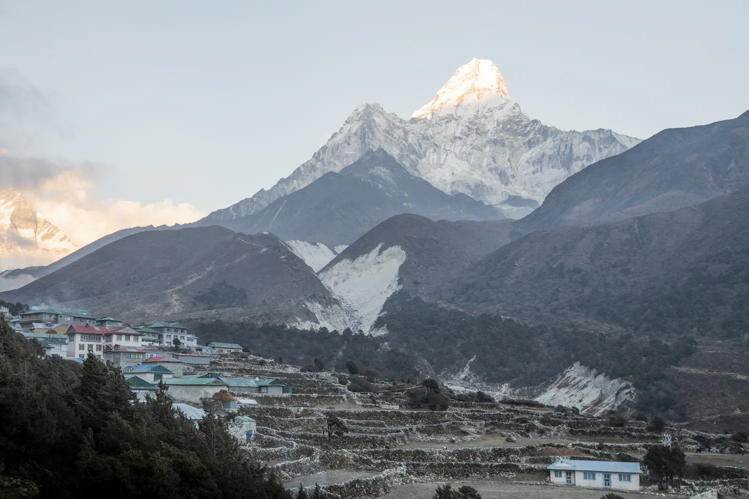 En liten fjellandsby med terrasserte steinmurer ligger i åssiden, mens den majestetiske, snødekte toppen av Ama Dablam fanger det varme sollyset i bakgrunnen.
