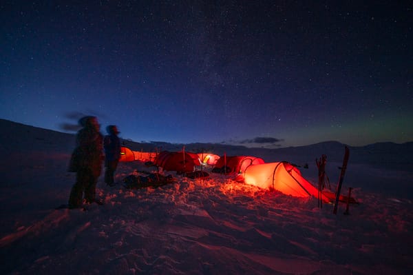 Oppsatte, lysende røde telt i snøen under en klar, stjerneklar nattehimmel, med to personer som ser opp mot stjernene.