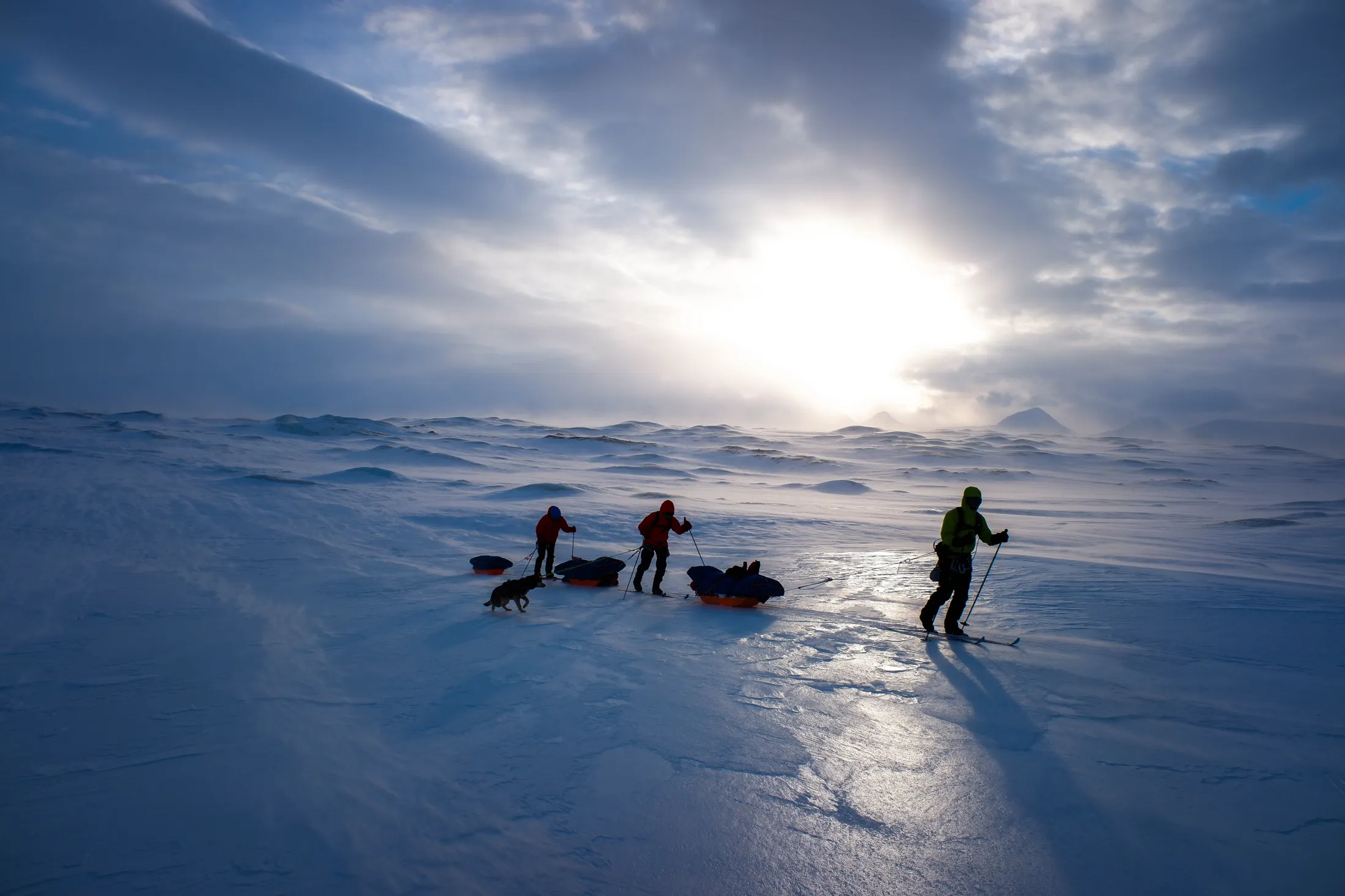 Tre skiløpere trekker lastede pulker over en enorm, islagt vidde i følge med en hund, silhuettert mot en dramatisk skyet himmel der solen bryter igjennom over fjerne fjell.