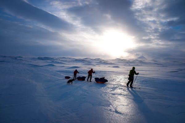 Tre skiløpere trekker lastede pulker over en enorm, islagt vidde i følge med en hund, silhuettert mot en dramatisk skyet himmel der solen bryter igjennom over fjerne fjell.