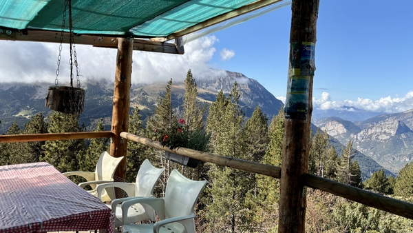 Utsikt fra en treterrasse med hvite stoler og et bord, med utsikt over en skog og fjerne fjell under en blå himmel.
