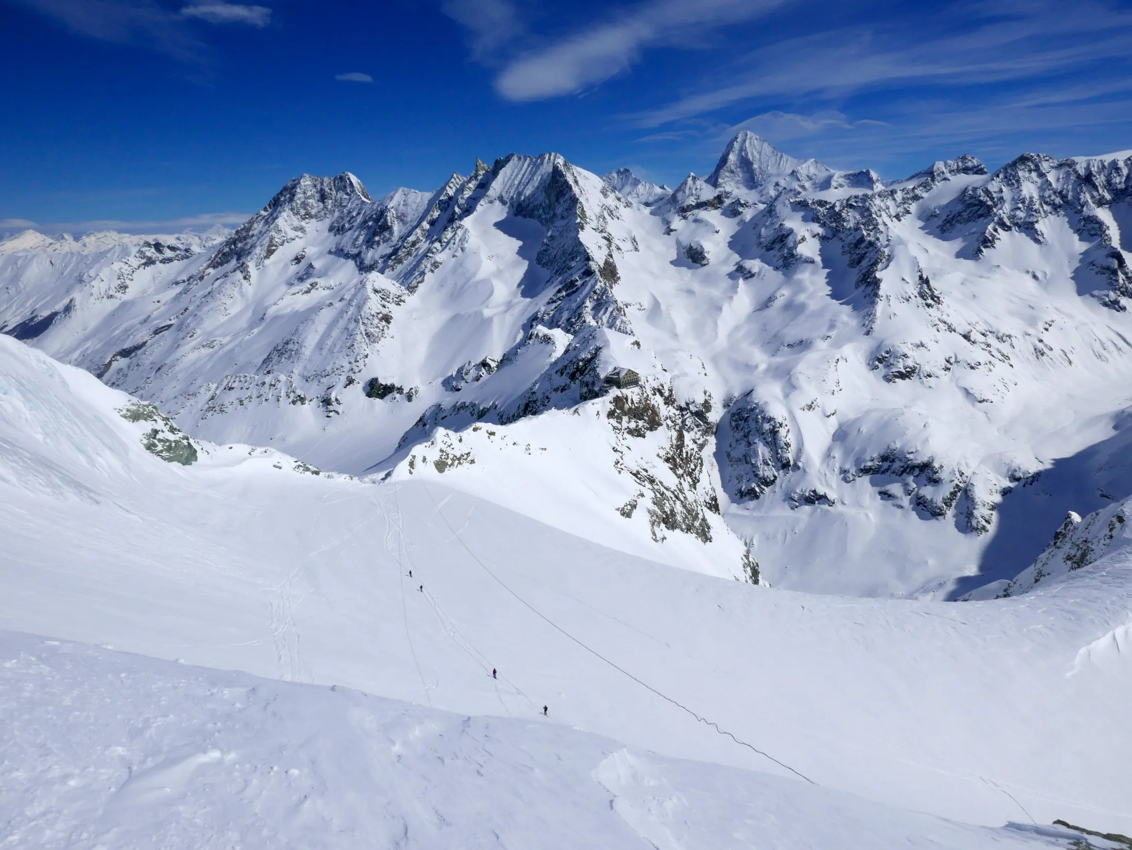 Små skikkelser av fjellklatrere går nedover et enormt, snødekt brebasseng omgitt av dramatiske alpine egger og spisse tinder under en dyp blå himmel.