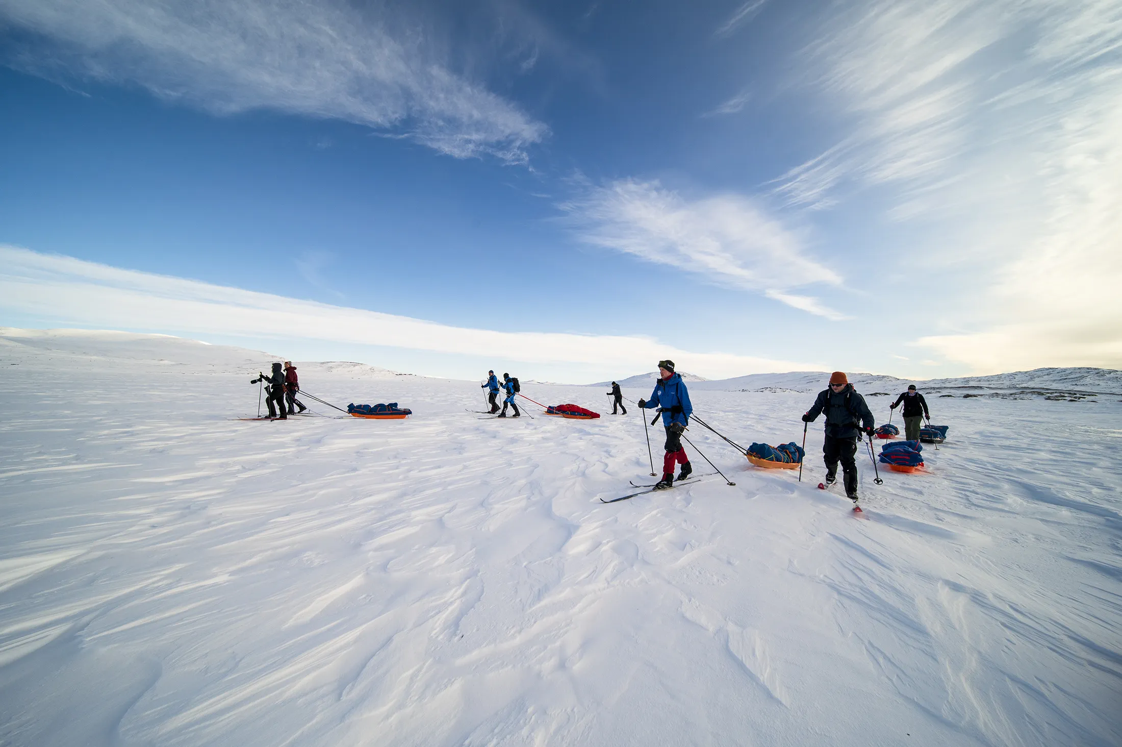 En gruppe skiløpere som trekker tunge pulker tvers over et vidstrakt, snødekket fjellandskap under en blå himmel.