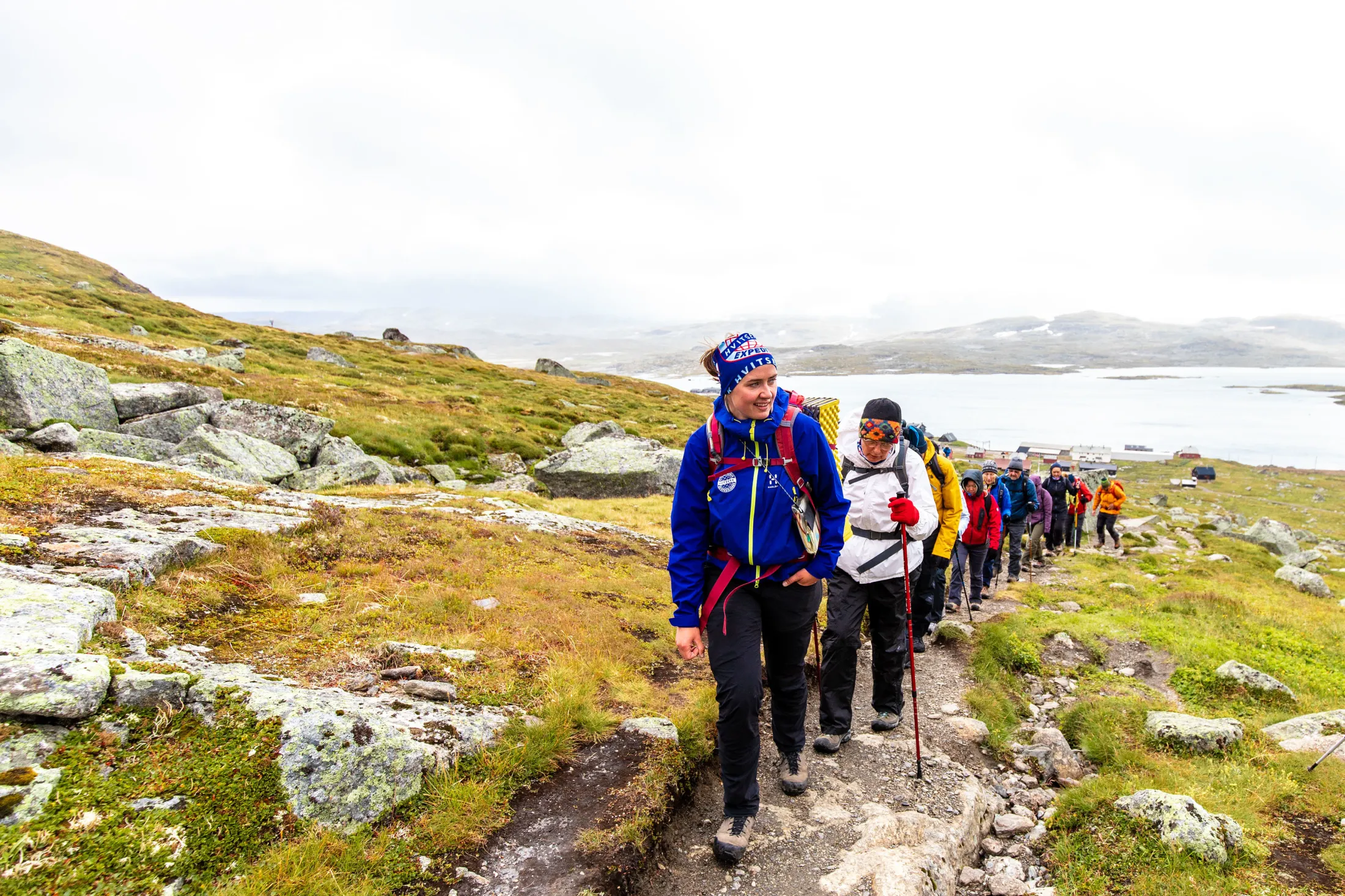 En stor gruppe turgåere går på en rekke nedover en steinete fjellsti på en overskyet dag, med et vann og fjerne bygninger i bakgrunnen.