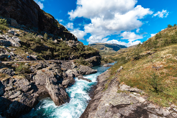 En brusende, turkis fjellelv renner gjennom et steinete gjel med syklister på en grusvei langs de bratte fjellsidene under en delvis skyet himmel.
