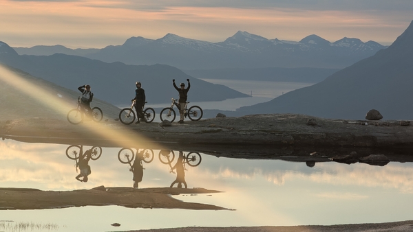 Tre syklister og skygger av dem reflektert i et vann med fjell i bakgrunnen.