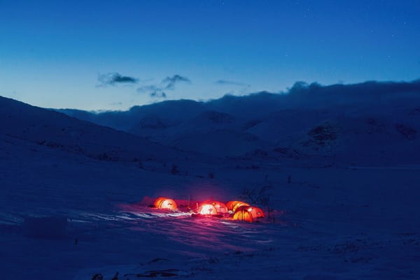 Et avstandsbilde av lysende røde telt slått opp i en snødekt fjelldal under en dypblå skumringshimmel.
