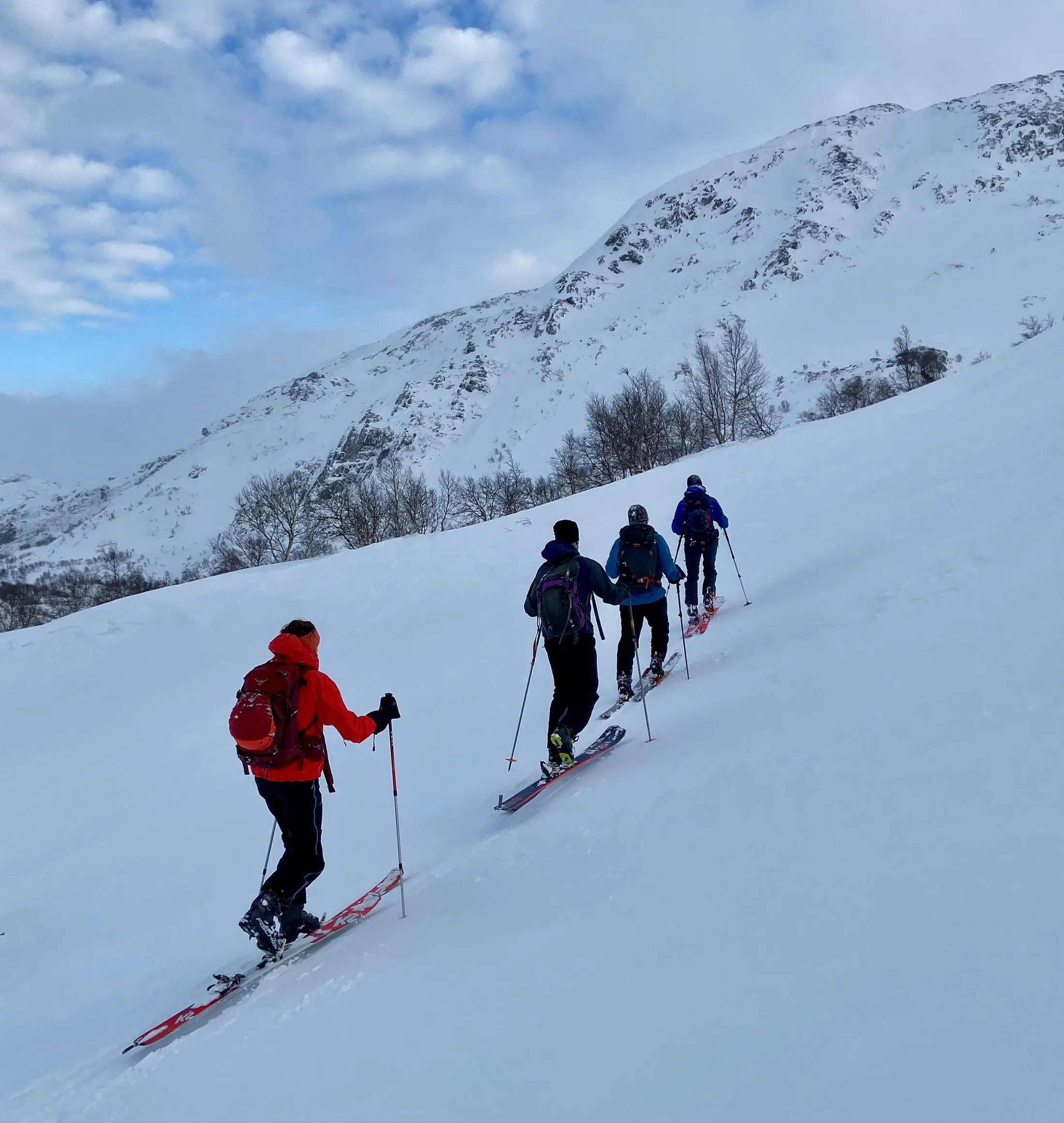 Fire toppturgåere som går oppover en snødekt fjellside under en skyet himmel.