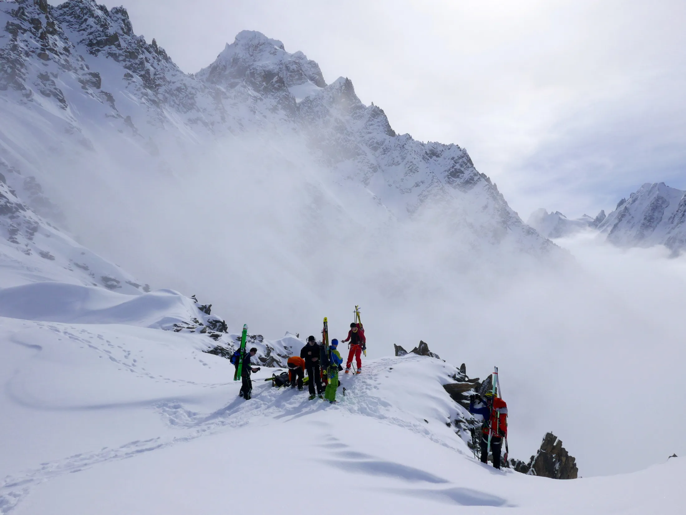 En gruppe skibestigere med utstyret sitt står på en snødekt fjellegg mens dramatiske skyer og tåke virvler rundt de omkringliggende, bratte tindene.