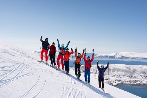 En gruppe skiløpere står på rad på en snødekt fjellkam med armene i været, med utsikt over havet på en klar dag.